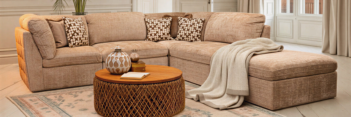 A beige upholstered sectional sofa with a chaise lounge, styled in a bright living room with patterned throw pillows, a round wooden coffee table, and a draped cream throw blanket.