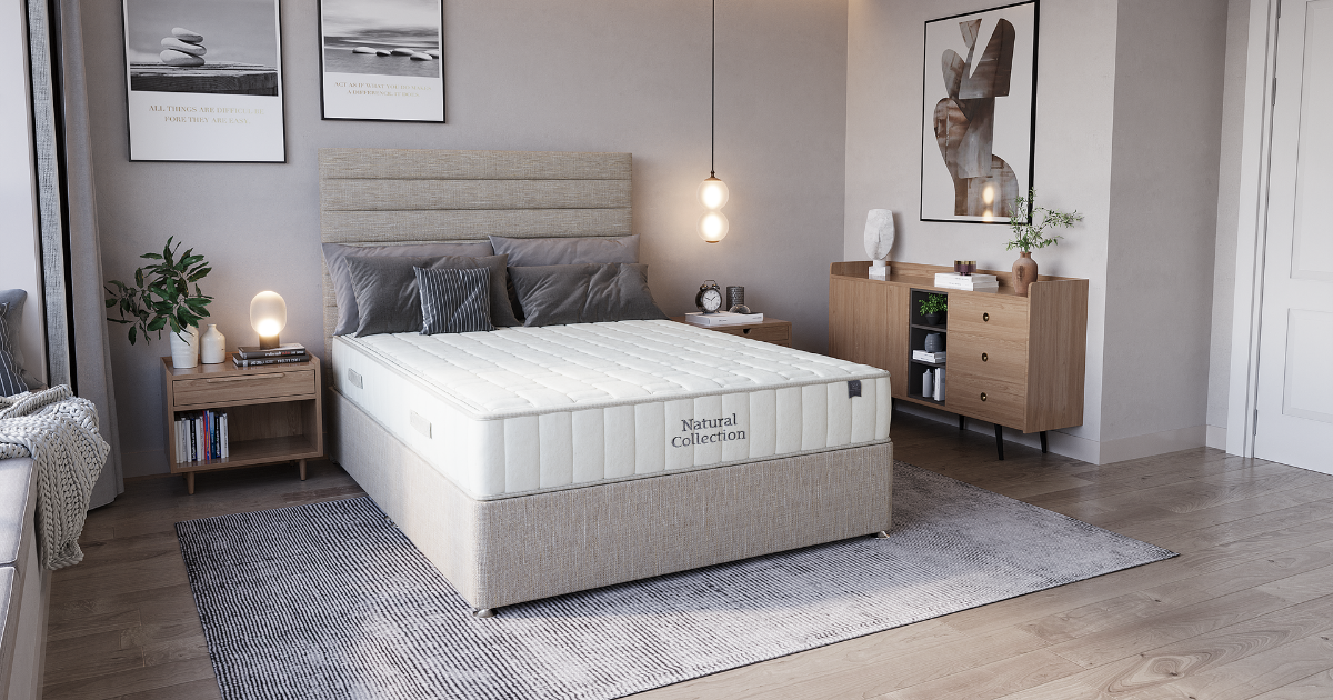 A king koil bed with a king koil mattress and a beige upholstered frame is shown in a styled bedroom setting with a gray rug, wooden nightstands, a dresser, and wall art.