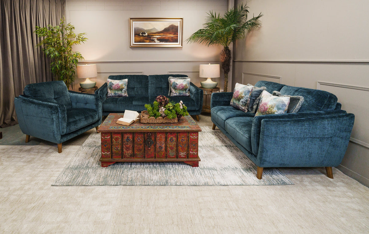 A staged living room featuring a matching three-seat sofa, armchair, and rectangular coffee table in deep teal velvet upholstery, arranged on a light rug with decorative pillows, lamps, plants, and a framed wall painting.