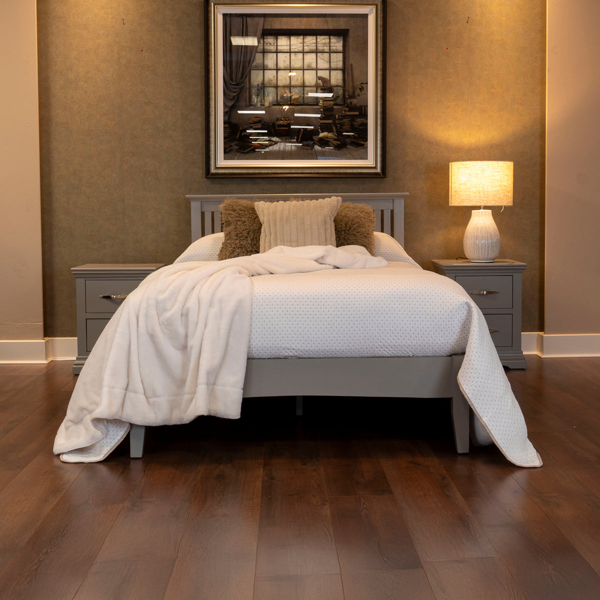 Grey upholstered bed frame with wooden legs, dressed with cream throws and cushions, styled bedroom with matching bedside tables, lamp, and framed art.